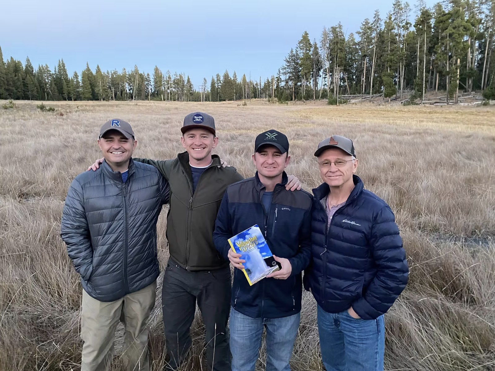 The family behind Yellowstone Explored, standing in a meadow at the edge of the park with a copy of the book
