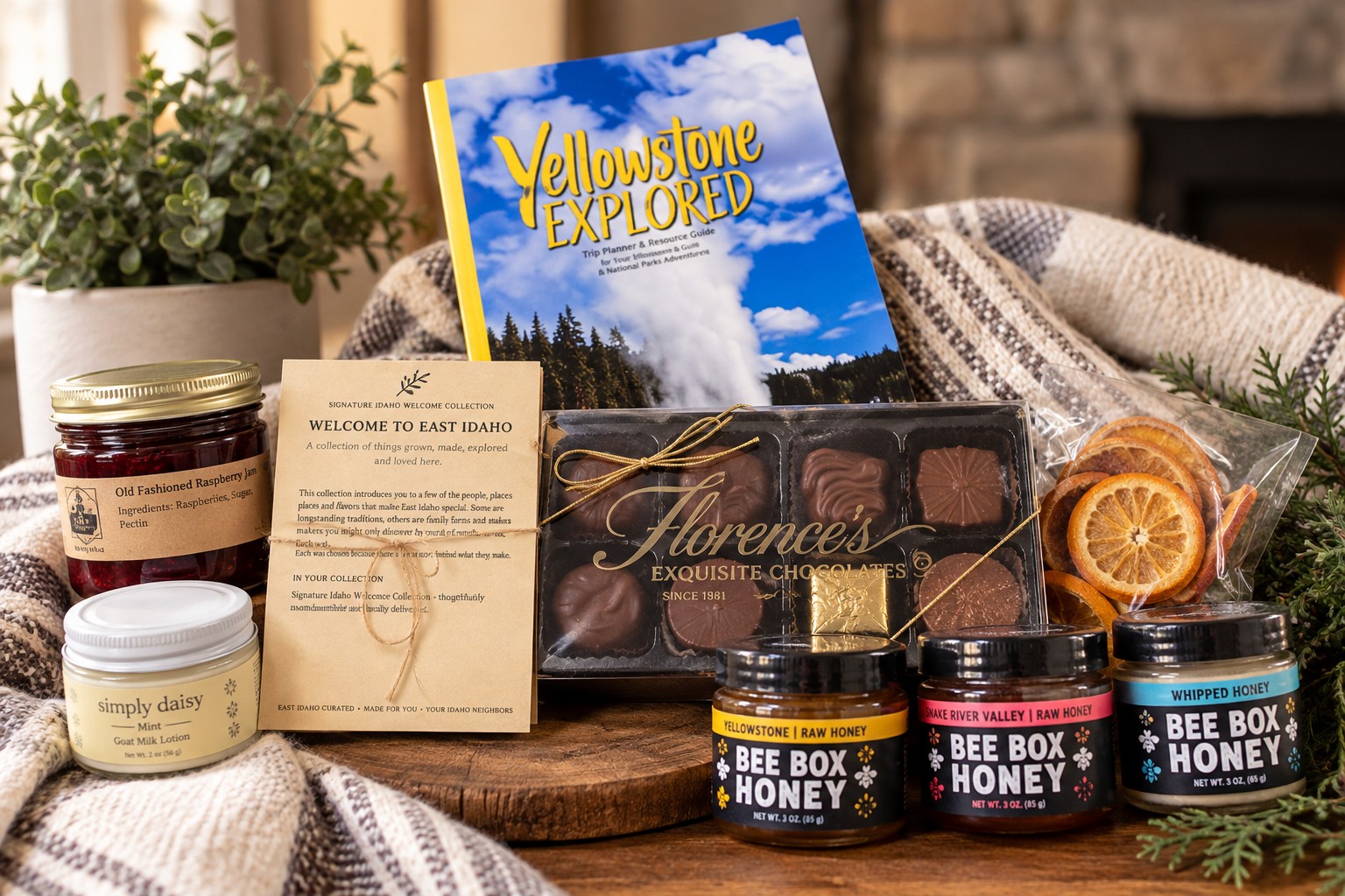 The Idaho Welcome Collection laid out before packing — Yellowstone Explored, raspberry jam, Simply Daisy goat milk lotion, the printed welcome card, Florence’s chocolates, the three Bee Box honeys and dried orange slices