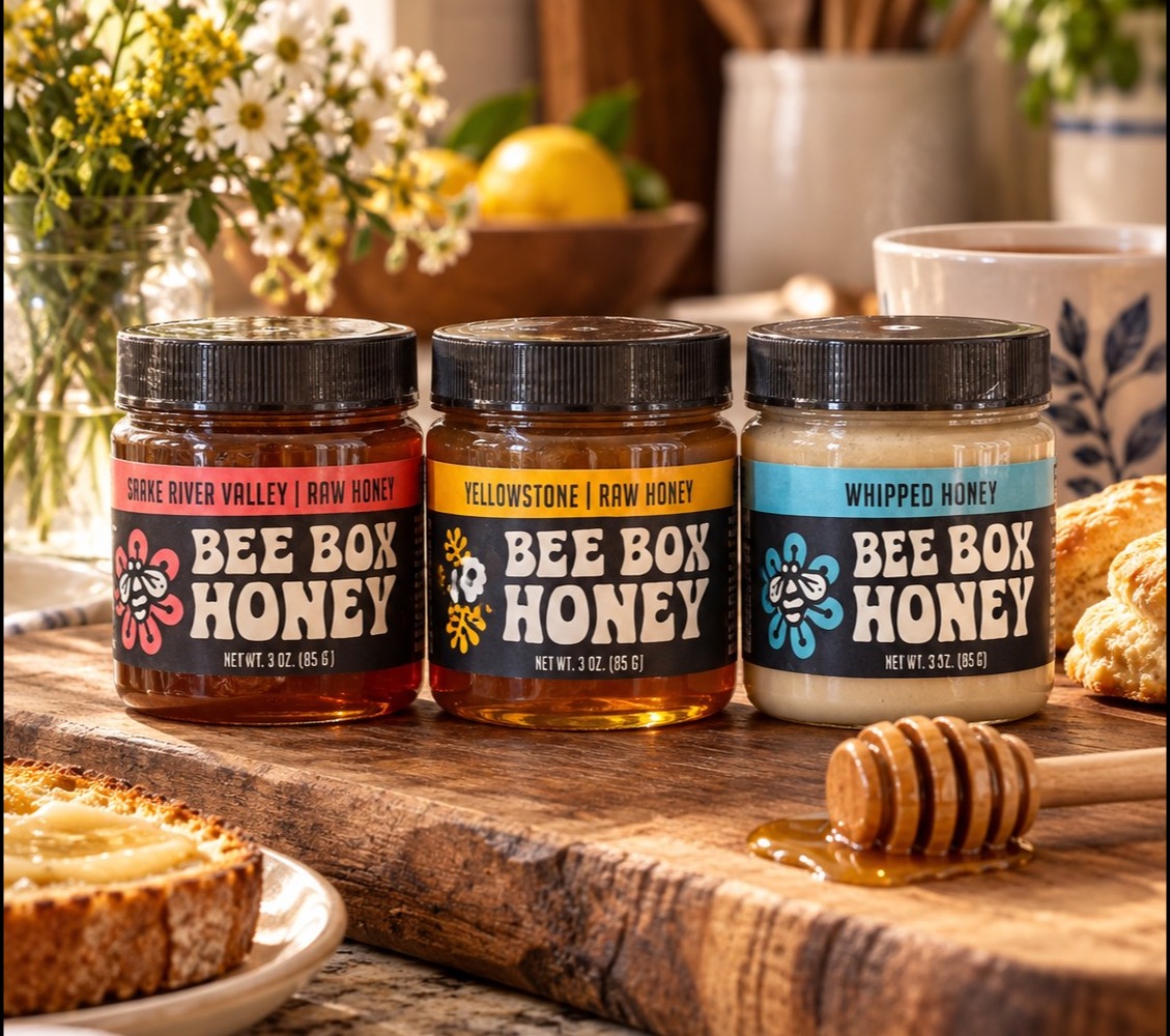 The three Bee Box honeys on a wooden board in a sunny kitchen, with wildflowers, toast and a honey dipper